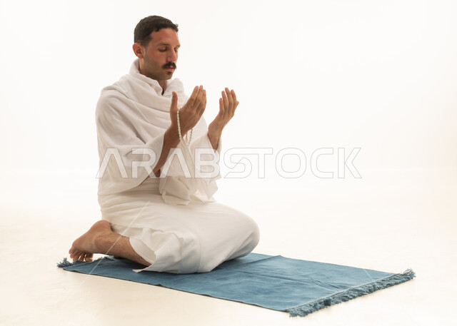 الركوع والسجود والخشوع لله، أذكار وأدعية وفرائض، رجل عربي سعودي خليجي يرتدي بملابس الإحرام  يمسك السبحة يجلس على السجادة و يؤدي فريضة الصلاة ، آداء صلاة التراويح فى شهر رمضان الكريم ، الدعاء والتضرع الى الله، خلفية بيضاء