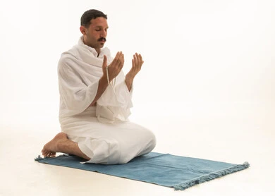 Bowing, prostration, and humility before God, remembrances, supplications, and obligations. An Arab Saudi Gulf man dressed in Ihram clothing holds a prayer bead, sits on a prayer rug, and performs the obligatory prayer, performing the Taraweeh prayer during the holy month of Ramadan, supplicating and beseeching God, with a white background.