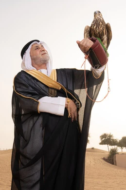 Raising birds and caring for their training and behavior improvement; the status of falcons in Emirati history; training a falcon of prey; an elderly Emirati Gulf Arab man wearing a white kandura, black bisht, shemagh, and agal stands in the desert holding a falcon on his hands; gestures of happiness and joy.