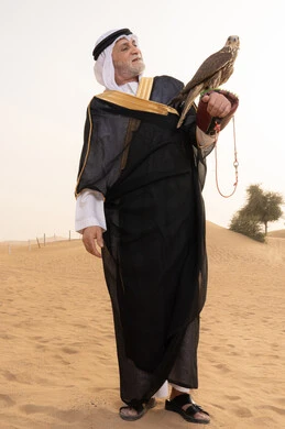 The art of taming birds of prey, developing birds' abilities through breeding, training the falcon, an elderly Emirati Gulf Arab man wearing a white kandura, black bisht, shemagh, and agal, standing in the desert holding a falcon on his hands, the place of falcons in Emirati history, gestures of happiness and joy