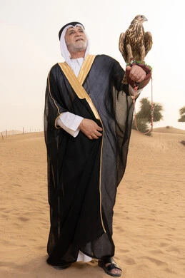 العناية بالطيور من خلال تربيتها وتدريبها، تدريب الصقر الجارح، رجل عربي خليجي إماراتي مسن يرتدي الكندورة البيضاء والبشت الأسود والشماغ مع العقال يقف فى الصحراء يحمل على يدية صقر, مكانة الصقور في التاريخ الاماراتي، ايماءات السعادة و السرور