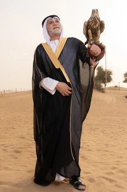 The care and training of birds, the training of the falcon, an elderly Emirati Gulf Arab man wearing a white kandura, black bisht, shemagh, and agal, standing in the desert holding a falcon on his hands, the place of falcons in Emirati history, gestures of happiness and joy