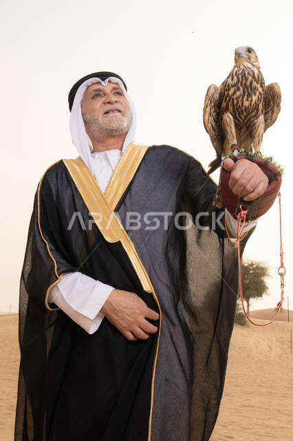 تدريب الصقر الجارح، تربية الطيور والاهتمام بتدريبها ورعايتها، رجل عربي خليجي إماراتي مسن يرتدي الكندورة البيضاء والبشت الأسود والشماغ مع العقال يقف فى الصحراء يحمل على يدية صقر, مكانة الصقور في التاريخ الاماراتي، ايماءات السعادة و السرور