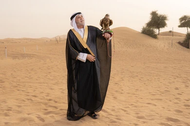 The interest in raising and training birds, authentic Emirati heritage, the concept of authenticity and preserving folklore, an elderly Emirati Gulf Arab man wearing a white kandura, black bisht, shemagh, and agal, standing in the desert holding a falcon in his hands and looking at it, the sport of falconry and a Bedouin atmosphere reflecting national identity