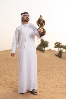 الاهتمام بتربيه الطيور البرية، تدريب الصقر الجارح, مكانة الصقور في التاريخ الاماراتي ، رجل عربي خليجي اماراتي يرتدي الكندورة و الغتره البيضاء يقف فى الصحراء يحمل على يده طائر الصقر