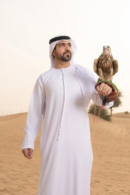 النظر الى شيء ما فى الاعلى، تدريب الصقر الجارح، الاهتمام بتربيه الطيور البرية, مكانة الصقور في التاريخ الاماراتي ، رجل عربي خليجي اماراتي يرتدي الكندورة و الغتره البيضاء يقف فى الصحراء يحمل على يده طائر الصقر