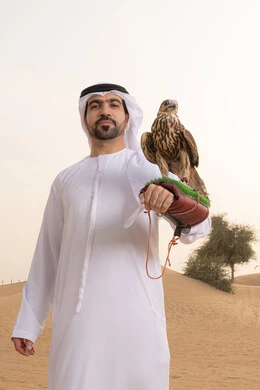النظر الى الكاميرا بتعابير السرور، تدريب الصقر الجارح، الاهتمام بتربيه الطيور البرية, مكانة الصقور في التاريخ الاماراتي ، رجل عربي خليجي اماراتي يرتدي الكندورة و الغتره البيضاء يقف فى الصحراء يحمل على يده طائر الصقر