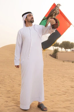 تدريب الصقر الجارح، الاهتمام بتربيه الطيور البرية, مكانة الصقور في التاريخ الاماراتي , استذكار اليوم الوطني الاماراتي 1971، ذكرى تأسيس الاتحاد 2 ديسمبر، رفع راية الوطن في يوم العلم 3 نوفمبر، رجل عربي خليجي اماراتي يرتدي الكندورة و الغتره البيضاء يحمل على يده طائر الصقر و علم الامارات بتعابير الفخر