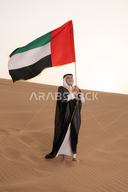 استذكار اليوم الوطني الاماراتي 1971، ذكرى تأسيس الاتحاد 2 ديسمبر، رفع راية الوطن في يوم العلم 3 نوفمبر، حب الوطن والاعتزاز به، رجل مسن عربي خليجي إماراتي يرتدي الكندورة والبشت مع الغترة البيضاء يمسك بيده علم الإمارات العربية المتحدة يقف في منطقة أثرية في الصحراء