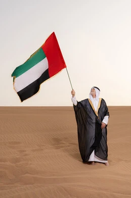 ذكرى تأسيس الاتحاد 2 ديسمبر، رفع راية الوطن في يوم العلم 3 نوفمبر، حب الوطن والاعتزاز به، استذكار اليوم الوطني الاماراتي 1971، رجل مسن عربي خليجي إماراتي يرتدي الكندورة والبشت مع الغترة البيضاء يمسك بيده علم الإمارات العربية المتحدة يقف في منطقة أثرية في الصحراء