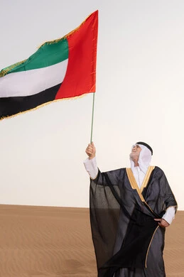 رفع راية الوطن في يوم العلم 3 نوفمبر، حب الوطن والاعتزاز به، ذكرى تأسيس الاتحاد 2 ديسمبر، استذكار اليوم الوطني الاماراتي 1971، رجل مسن عربي خليجي إماراتي يرتدي الكندورة والبشت مع الغترة البيضاء يمسك بيده علم الإمارات العربية المتحدة يقف في منطقة أثرية في الصحراء