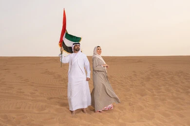 العيد الوطني الإماراتي، رجل عربي خليجي اماراتي يرتدي الكندورة والغترة يقف فى الصحراء برفقة زوجته يمسك بيدية علم الامارات، ايماءات السعادة والفخر والاعتزاز، اليوم الوطني الإماراتي 2 ديسمبر، ذكرى استقلال دولة الإمارات العربية المتحدة، علم وراية الوطن، حب الوطن والانتماء اليه