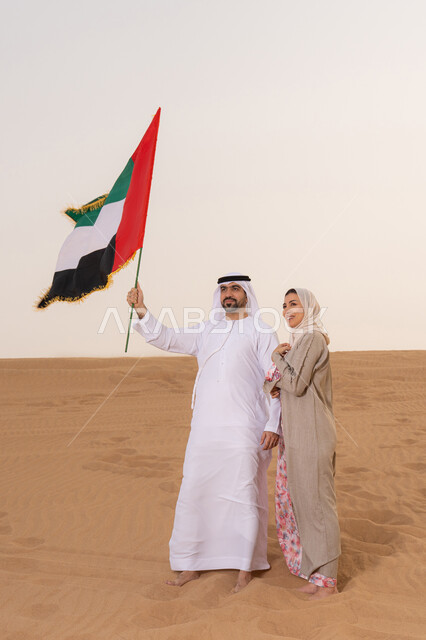 اليوم الوطني الإماراتي 2 ديسمبر، ذكرى استقلال دولة الإمارات العربية المتحدة، علم وراية الوطن، حب الوطن والانتماء اليه، ايماءات السعادة والفخر والاعتزاز، رجل عربي خليجي اماراتي يرتدي الكندورة والغترة يقف فى الصحراء برفقة زوجته يمسك بيدية علم الامارات