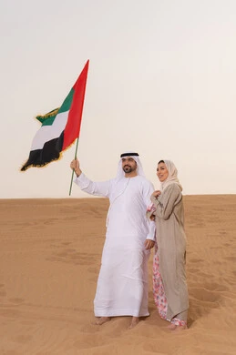 اليوم الوطني الإماراتي 2 ديسمبر، ذكرى استقلال دولة الإمارات العربية المتحدة، علم وراية الوطن، حب الوطن والانتماء اليه، ايماءات السعادة والفخر والاعتزاز، رجل عربي خليجي اماراتي يرتدي الكندورة والغترة يقف فى الصحراء برفقة زوجته يمسك بيدية علم الامارات