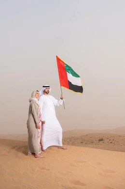 ذكرى استقلال دولة الإمارات العربية المتحدة، علم وراية الوطن، حب الوطن والانتماء اليه، ايماءات السعادة والفخر والاعتزاز، اليوم الوطني الإماراتي 2 ديسمبر، رجل عربي خليجي اماراتي يرتدي الكندورة والغترة يقف فى الصحراء برفقة زوجته يمسك بيدية علم الامارات