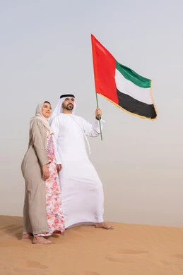 علم وراية الوطن، حب الوطن والانتماء اليه، ايماءات السعادة والفخر والاعتزاز، ذكرى استقلال دولة الإمارات العربية المتحدة، اليوم الوطني الإماراتي 2 ديسمبر، رجل عربي خليجي اماراتي يرتدي الكندورة والغترة يقف فى الصحراء برفقة زوجته يمسك بيدية علم الامارات