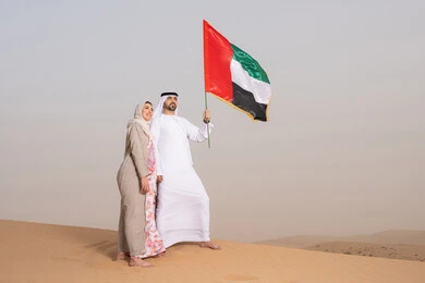 حب الوطن والانتماء اليه، ايماءات السعادة والفخر والاعتزاز، علم وراية الوطن، ذكرى استقلال دولة الإمارات العربية المتحدة، اليوم الوطني الإماراتي 2 ديسمبر، رجل عربي خليجي اماراتي يرتدي الكندورة والغترة يقف فى الصحراء برفقة زوجته يمسك بيدية علم الامارات