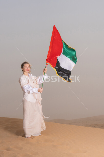 ذكرى تأسيس الاتحاد 2 ديسمبر، استذكار اليوم الوطني الاماراتي 1971، إمراة عربية خليجية إماراتية ترتدي العباءة تمسك بيدها علم الإمارات العربية المتحدة تقف في الصحراء، يوم العلم 3 نوفمبر، حب الوطن والاعتزاز به
