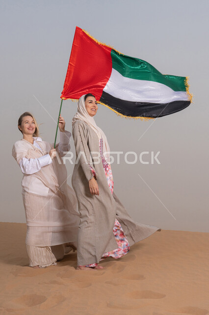 إحياء اليوم الوطني الإماراتي 2 ديسمبر، الاحتفال بذكرى استقلال دولة الإمارات العربية المتحدة، امراة عربية خليجية اماراتية ترتدي العباءة تقف فى الصحراء برفقة صديقتها تمسك علم الإمارات، ايماءات السعادة والفخر والاعتزاز، مفهوم حب الوطن والانتماء اليه، النظر الى شيء ما