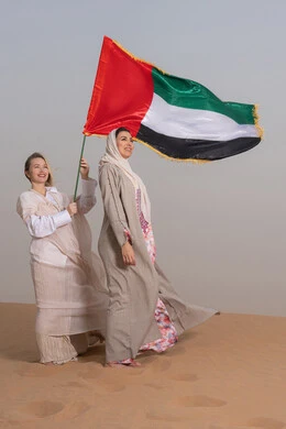 Commemorating UAE National Day on December 2nd, the anniversary of the independence of the United Arab Emirates, an Emirati woman wearing an abaya stands in the desert with her friend, holding the UAE flag. Gestures of happiness, pride, and honor; the concept of patriotism and belonging; looking at something