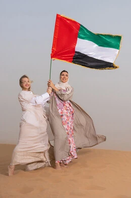 Commemorating the UAE National Day 1971, Union Day, December 2nd; the concept of patriotism and belonging; celebrating national holidays and occasions; two Gulf Arab women wearing abayas stand in the desert holding the UAE flag; Flag Day commemoration, November 3rd.
