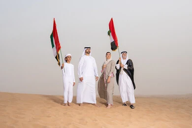 أجواء تراثية تعكس الأصالة والانتماء الوطني، مفهوم الفخر والوحدة الوطنية،الاحتفال بالهوية الوطنية والانتماء, عائلة عربية خليجية إماراتية ترتدي الزي التقليدي تقف في الصحراء وترفع علم دولة الإمارات العربية المتحدة، النظر الى شيء ما