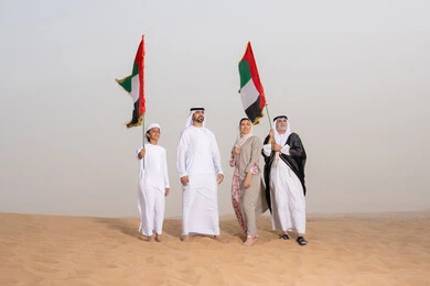 مفهوم الفخر والوحدة الوطنية،الاحتفال بالهوية الوطنية والانتماء, عائلة عربية خليجية إماراتية ترتدي الزي التقليدي تقف في الصحراء وترفع علم دولة الإمارات العربية المتحدة، النظر الى شيء ما, أجواء تراثية تعكس الأصالة والانتماء الوطني