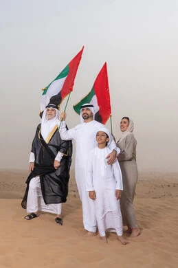 Celebrating national identity and belonging, the concept of pride and national unity; an Emirati Gulf Arab family wearing traditional dress stands in the desert and raises the flag of the United Arab Emirates, gazing at something; a heritage atmosphere reflecting authenticity and national belonging.
