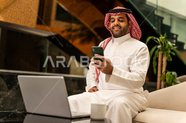 رجل خليجي سعودي بداخل احدى الفنادق السياحية ، يحمل الهاتف المحمول ، إستخدام جهاز الحاسوب المحمول لإنجاز المهام، مفهوم التكنولوجيا وإستخدام أجهزة تقنية حديثة، العمل عن بعد ، مهن و وظائف ، جلسة عمل، مفهوم إدارة الأعمال، سياحة وفندقة، التسويق الإلكتروني