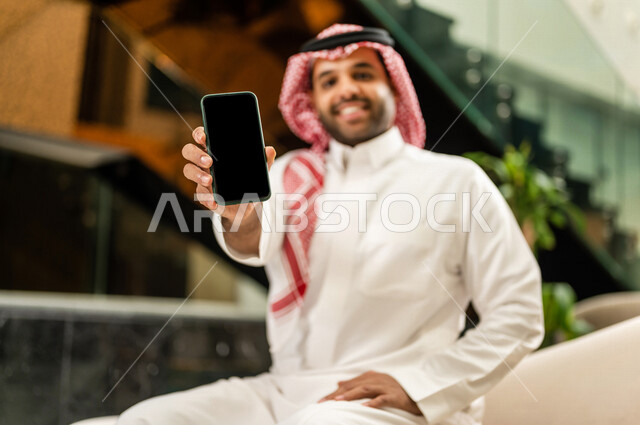 رجل خليجي سعودي بداخل احدى الفنادق السياحية ، يمسك بيده الهاتف المحمول ، يعرض شاشة سوداء ، مفهوم إستخدام أجهزة تقنية حديثة، عرض خدمات وتطبيقات عبر الجوال ، التسويق الرقمي الإلكتروني ، مفهوم الإعلانات، رحلة عمل، سياحة وفندقة ، جلسة عمل، سياحة وسفر
