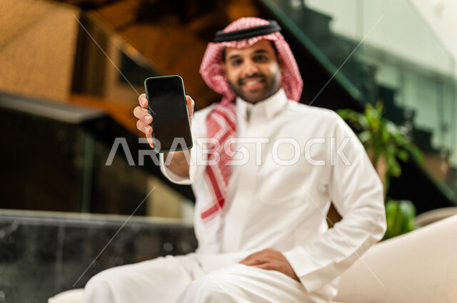 رجل خليجي سعودي بداخل احدى الفنادق السياحية ، يمسك بيده الهاتف المحمول ، يعرض شاشة سوداء ، مفهوم إستخدام أجهزة تقنية حديثة، عرض خدمات وتطبيقات عبر الجوال ، التسويق الرقمي الإلكتروني ، مفهوم الإعلانات، رحلة عمل، سياحة وفندقة ، جلسة عمل، سياحة وسفر