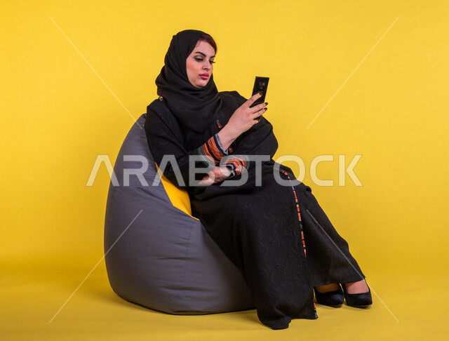 Saudi Gulf Arab woman sitting on the sofa, browsing the mobile phone, chatting and conversations, communicating with friends, video calls, reading from the mobile phone, having a good and quiet time, having fun, yellow background