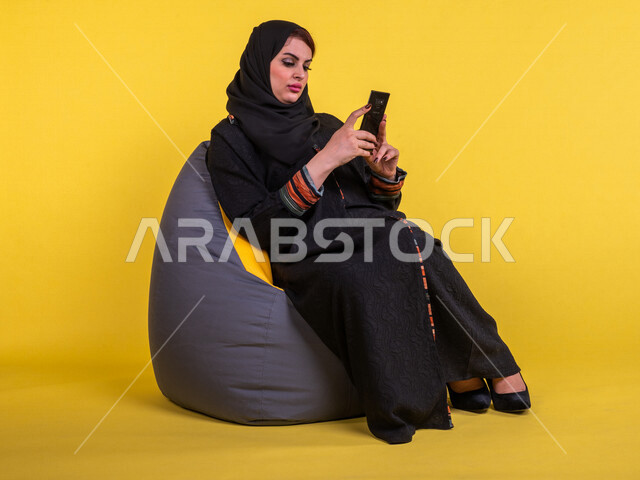 إمرأه عربيه سعوديه خليجيه تجلس على الأريكه, تصفح الهاتف المحمول, دردشه ومحادثات, التواصل مع الأصدقاء, مكالمات فيديو, القراءه من الهاتف المحمول, قضاء وقت ممتع وهادئ, تسليه وترفيه, خلفيه صفراء