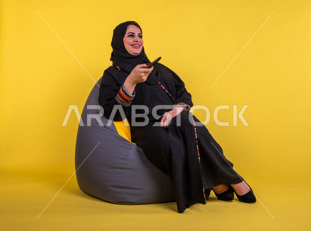 إمرأه عربيه سعوديه خليجيه تجلس على الأريكه وتشاهد التلفاز, استخدام عصا التحكم, التحكم بالتلفاز عن بعد, قضاء وقت ممتع , تسليه وترفيه, ايماءات وجه تدل على السعاده والاستمتاع, خلفيه صفراء