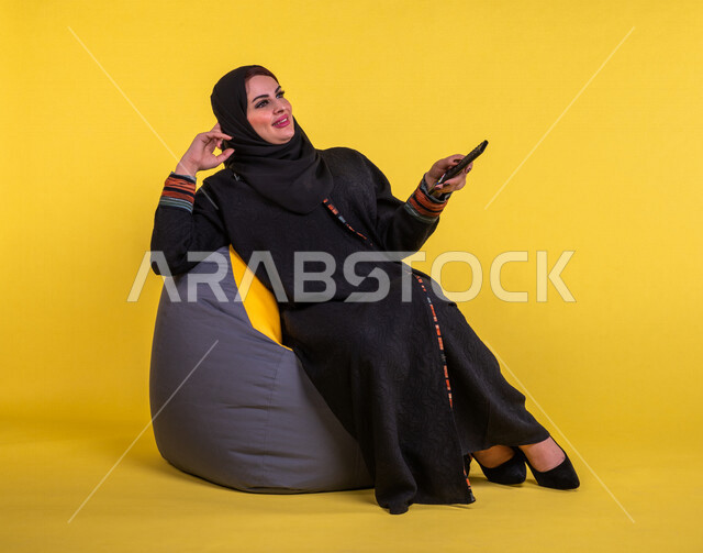 إمرأه عربيه سعوديه خليجيه تجلس على الأريكه وتشاهد التلفاز, استخدام عصا التحكم, التحكم بالتلفاز عن بعد, قضاء وقت ممتع , تسليه وترفيه, ايماءات وجه تدل على السعاده والاستمتاع, خلفيه صفراء