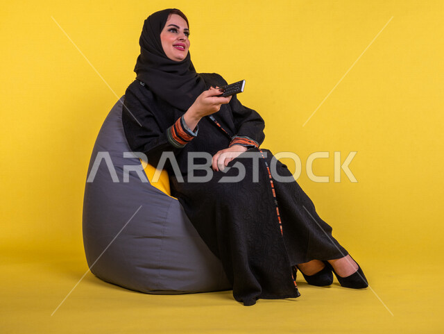 إمرأه عربيه سعوديه خليجيه تجلس على الأريكه وتشاهد التلفاز, استخدام عصا التحكم, التحكم بالتلفاز عن بعد, قضاء وقت ممتع , تسليه وترفيه, ايماءات وجه تدل على السعاده والاستمتاع, خلفيه صفراء