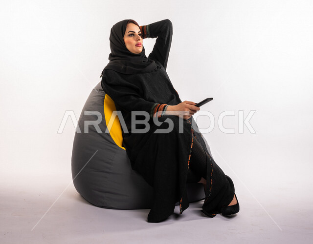 إمرأه عربيه سعوديه خليجيه تجلس على الأريكه وتشاهد التلفاز, استخدام عصا التحكم عن بعد, التحكم بالتلفاز عن بعد, قضاء وقت ممتع , تسليه وترفيه, ايماءات وجه تدل على السعاده والاستمتاع, خلفيه صفراء