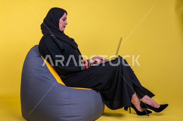 A Saudi Arabian Gulf woman sitting on a comfortable sofa, completing work tasks via the computer, managing projects remotely, playing on the laptop, making video calls over the Internet, having fun and having a good time