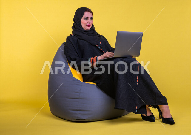 إمرأه عربيه سعوديه خليجيه تجلس على أريكه مريحه, انجاز مهام العمل عبر الحاسوب, إدارة المشاريع عن بعد, اللعب على اللابتوب, إجراء مكالمات فيديو عبر الإنترنت, تسليه وترفيه, قضاء وقت ممتع, خلفيه صفراء