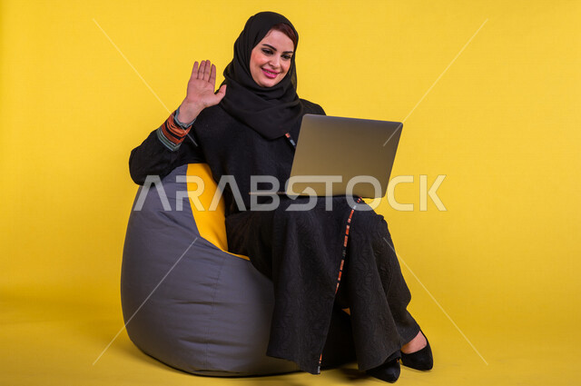 A Saudi Arabian Gulf woman sitting on a comfortable sofa, completing work tasks via the computer, managing projects remotely, playing on the laptop, making video calls over the Internet, having fun and having a good time