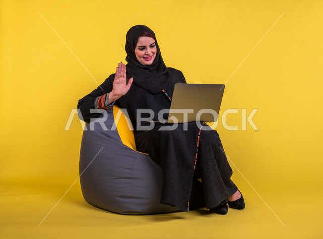 إمرأه عربيه سعوديه خليجيه تجلس على أريكه مريحه, انجاز مهام العمل عبر الحاسوب, إدارة المشاريع عن بعد, اللعب على اللابتوب, إجراء مكالمات فيديو عبر الإنترنت, تسليه وترفيه, قضاء وقت ممتع, خلفيه صفراء