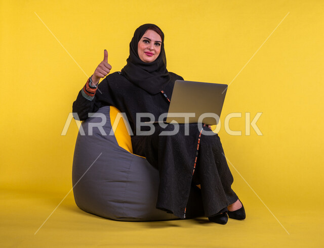 إمرأه عربيه سعوديه خليجيه تجلس على أريكه مريحه, انجاز مهام العمل عبر الحاسوب, إدارة المشاريع عن بعد, اللعب على اللابتوب, إجراء مكالمات فيديو عبر الإنترنت, تسليه وترفيه, قضاء وقت ممتع, خلفيه صفراء