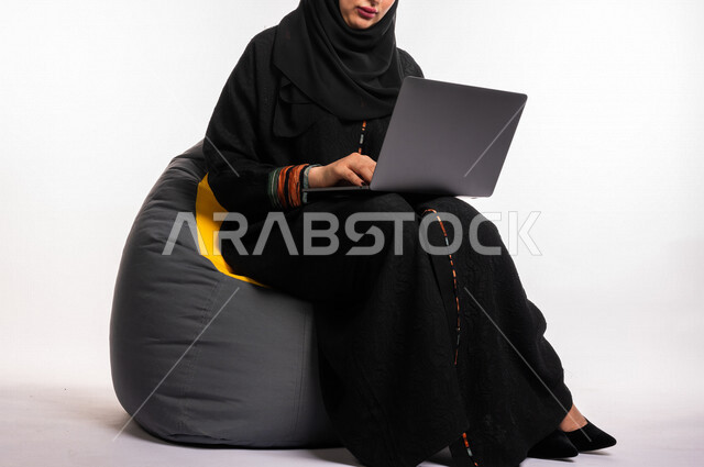 Portrait of a Saudi Arabian Gulf woman sitting on a comfortable sofa, accomplishing work tasks via the computer, managing projects remotely, playing on the laptop, making video calls over the Internet, watching a movie on the mobile device, entertainment,