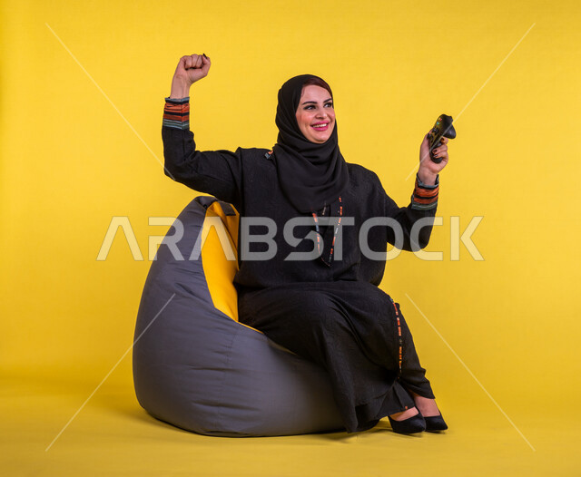 إمرأه عربيه سعوديه خليجيه تجلس على أريكه مريحه, اللعب بألعاب البلايستيشن,  استخدام يد التحكم عن بعد, ايماءات وجه ويدين تدل على التفاعل والإندماج مع اللعبه, تسليه وترفيه, قضاء وقت ممتع مع الألعاب الاكترونيه