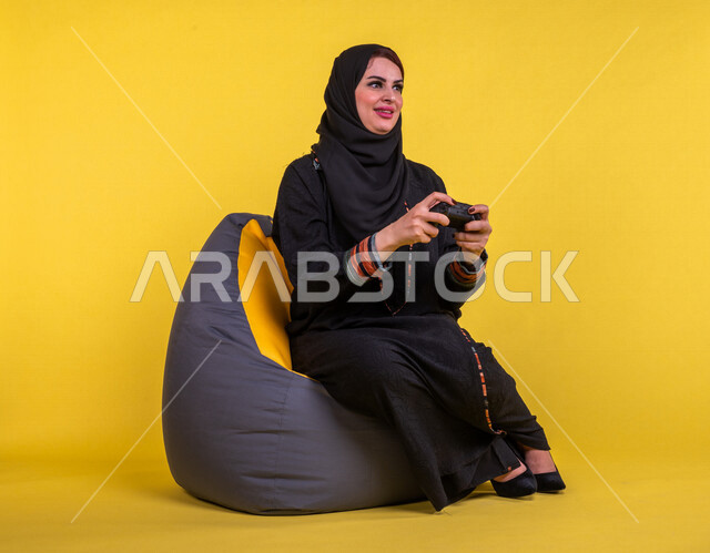 إمرأه عربيه سعوديه خليجيه تجلس على أريكه مريحه, اللعب بألعاب البلايستيشن,  استخدام يد التحكم عن بعد, ايماءات وجه ويدين تدل على التفاعل والإندماج مع اللعبه, تسليه وترفيه, قضاء وقت ممتع مع الألعاب الاكترونيه