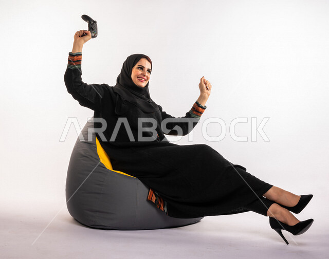 بورتريه لإمرأه عربيه سعوديه خليجيه تجلس على أريكه مريحه, اللعب بألعاب البلايستيشن,  استخدام يد التحكم عن بعد, ايماءات وجه ويدين تدل على التفاعل والإندماج مع اللعبه, تسليه وترفيه, قضاء وقت ممتع مع الألعاب الاكترونيه