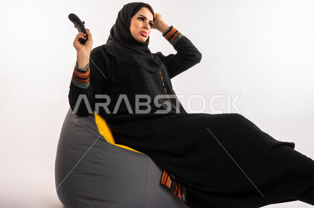 بورتريه لإمرأه عربيه سعوديه خليجيه تجلس على أريكه مريحه, اللعب بألعاب البلايستيشن,  استخدام يد التحكم عن بعد, ايماءات وجه ويدين تدل على التفاعل والإندماج مع اللعبه, تسليه وترفيه, قضاء وقت ممتع مع الألعاب الاكترونيه