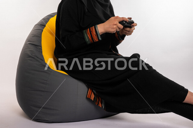 بورتريه لإمرأه عربيه سعوديه خليجيه تجلس على أريكه مريحه, اللعب بألعاب البلايستيشن,  استخدام يد التحكم عن بعد, ايماءات وجه ويدين تدل على التفاعل والإندماج مع اللعبه, تسليه وترفيه, قضاء وقت ممتع مع الألعاب الاكترونيه