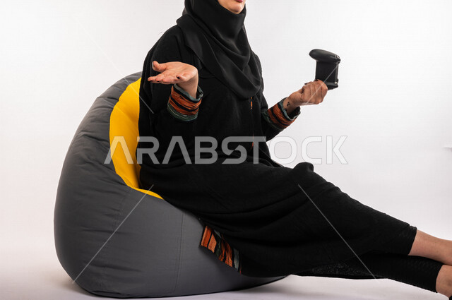 بورتريه لإمرأه عربيه سعوديه خليجيه تجلس على أريكه مريحه, اللعب بألعاب البلايستيشن,  استخدام يد التحكم عن بعد, ايماءات وجه ويدين تدل على التفاعل والإندماج مع اللعبه, تسليه وترفيه, قضاء وقت ممتع مع الألعاب الاكترونيه