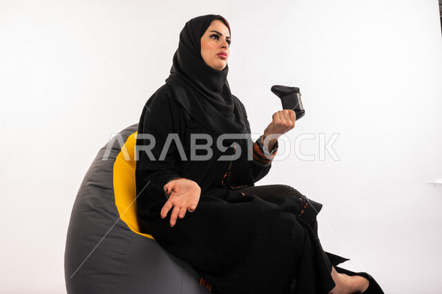 بورتريه لإمرأه عربيه سعوديه خليجيه تجلس على أريكه مريحه, اللعب بألعاب البلايستيشن,  استخدام يد التحكم عن بعد, ايماءات وجه ويدين تدل على التفاعل والإندماج مع اللعبه, تسليه وترفيه, قضاء وقت ممتع مع الألعاب الاكترونيه